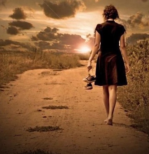 A imagem mostra uma mulher de costas, descalça, caminha numa estrada de terra , no por-do-sol, segurando um par de tênis na mão esquerda.