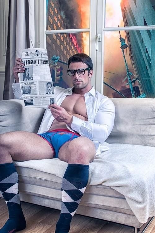 homem jovem, musculoso, sentado num sofá , rente a uma janela de vidro, ele lê uma pagina de jornal, usando apenas uma cueca azul, camisa social branca, e meias de cores quadriculadas.
