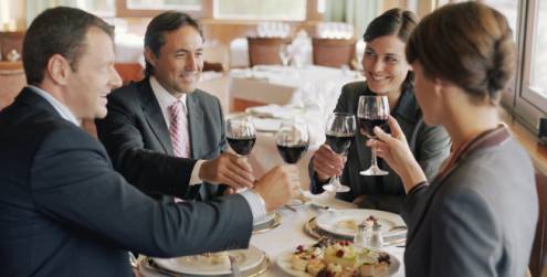 dois homens de terno e duas mulheres vestidas também com roupas sociais estão à mesa e brindam com taças de vinho