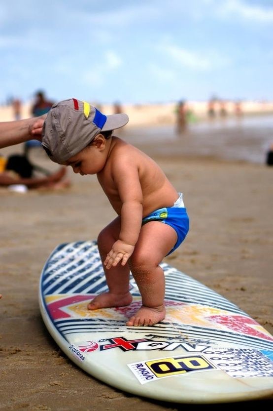 criança de dois anos , em cima de uma prancha de surf sobre a areia, ela usa boné ao contrário e está se equilibrando.