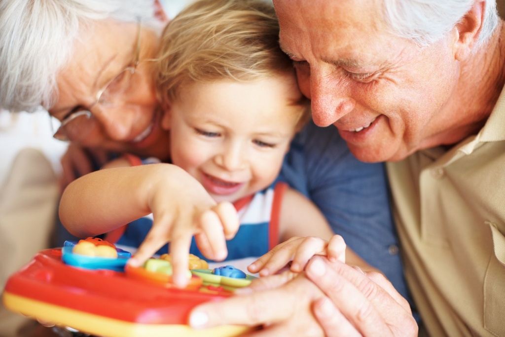 A imagem mostra um avô e uma avó de cabelos brancos, como rostos colados em um menino de 1 ano loiro com olhos azuis mexendo em um brinquedo de plástico. Mostra bem a ternura entre avós e netos