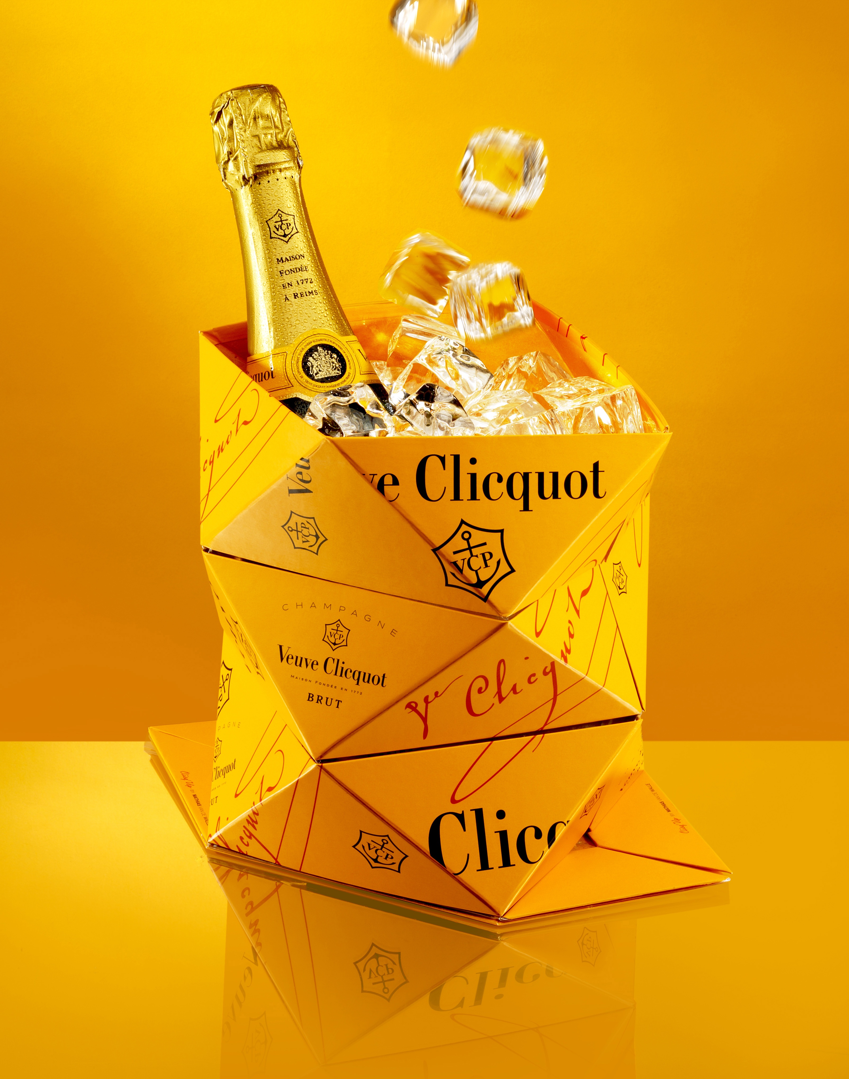 Num fundo todo amarelo ouro, temos um Balde de gelo, em forma de dobradura triangular, na cor amarelo ouro, classico da Champanhe Veuve Clicquot. Dentro dele um garrafa da champanhe fechada e muito gelo.