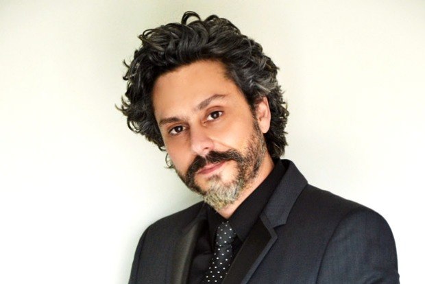 Ator Alexandre Nero, com imagem do personagem da novela "Império" da Rede Globo, está em imagem lateral, com cabelos, barba e bigode pretos grisalhos, veste camisa e paletó na cor preta, e grava preta com pequenas bolas brancas.