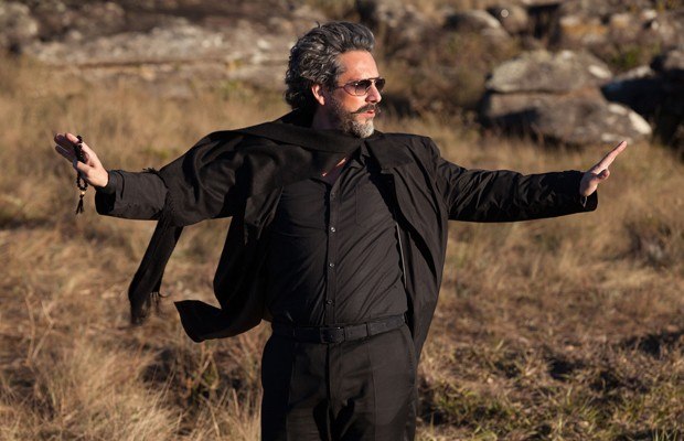 ator Alexandre Nero, como personagem José Alfredo da novela Império, está no Monte de Roraima, veste calça, camisa e jaqueta na cor preta, está com os braços abertos em ambas direções, usa óculos escuros e seus cabelos grisalhos escuros.