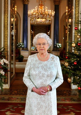 Rainha Elizabeth , usando um vestido azul claro, com as mãos entrelaçadas junto a região do baixo abdomen, parada no corredor do Palácio em Londres, ladeada, a esquerda por um arranjo de natal e a direita por uma árvore de natal , grande, com bolas vermelhas e amarelas .
