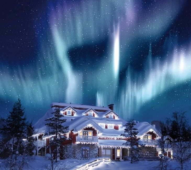 Foto noturna, de uma casa de vários andares na neve. Telhados e tudo que a rodeia está branco com a neve. Ao fundo o céu escuro é iluminado por um impressionante clarão verde e violeta de intensidade fulgurante - a famosa aurora boreal.