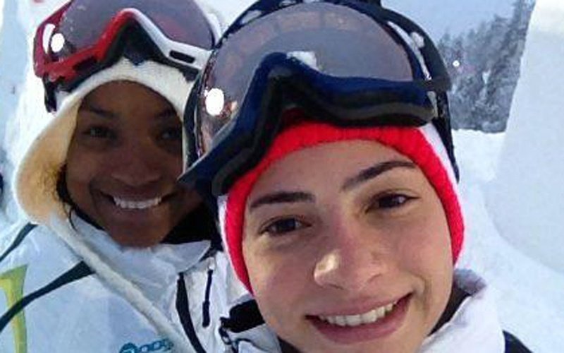 Lais de Souza, atleta brasileira, em imagem do rosto, está de sorrindo, com uma proteção na cabeça e um óculos para neve, ao fundo um outro esquiador também sorri.