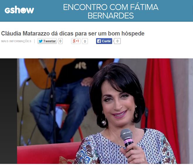 Claudia Matarazzo no programa Encontro com Fátima Bernardes, sentada no sofá em tom de vermelho, ela veste um blusa na cor azul acinzentado com pequenos detalhes, florzinhas, ela está com o microfone na mão direita, a frente junto a região do pescoço.