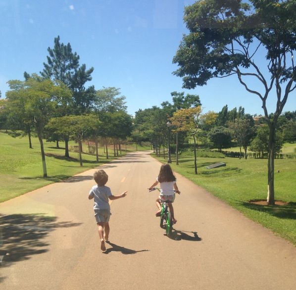 Numa manhã de sol, céu azul, numa rua de um parque, temos uma imagem de costas, de um menino corre junto a uma menina que anda de bicicleta.