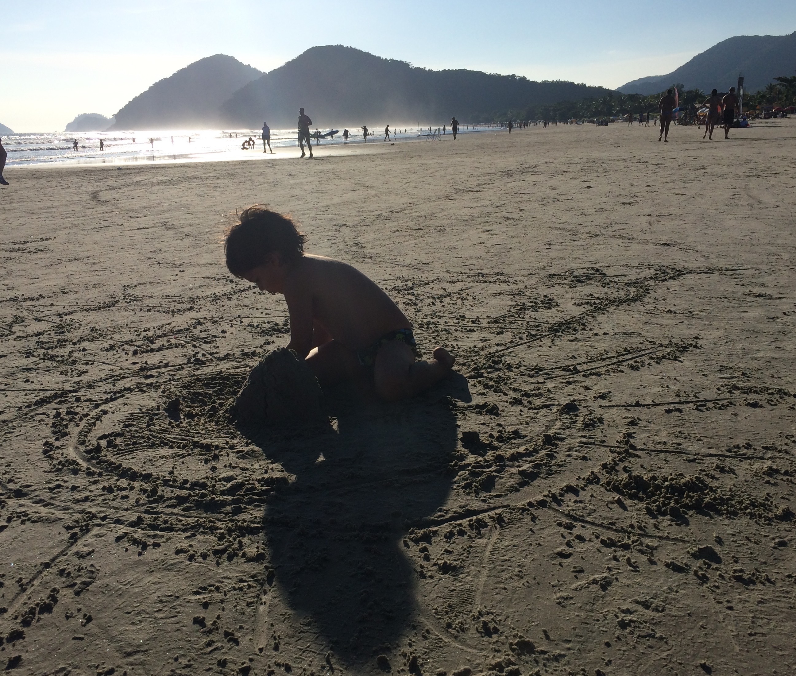 Num dia de sol, criança pequena, brinca na praia, fazendo montes de areia. Ele aparece em primeiro plano e ao fundo , muito longe, algumas outras pessoas transitam na praia.