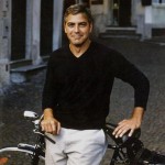 Ator George Clooney, durante as filmagens, está usando uma camiseta com decote em "V" na cor preta, manga longa e calça social na cor branca. Ele está sentado numa bicicleta e faz um sorriso contido e olha fixo para a câmera.