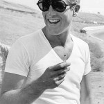 Ator Paul Newman, veste camiseta com decote "V", ma cor branca, e sobre ela uma jaqueta jeans. Ele usa óculos escuros olha para o horizonte. Segura na mão direita um cigarro aceso.