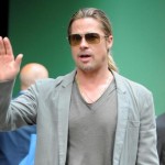Ator Brad Pitt, veste camiseta com decote "V", ma cor verde oliva e sobre ela um blaser na cor verde oliva mais clara. Ele usa óculos escuros olha para o horizonte e acena com a mão direita.