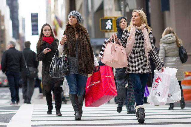 Varias mulheres andando numa rua e todas estão com muitas sacolas de compras de diversas cores.