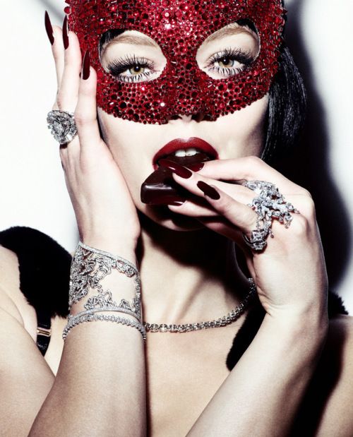imagem de um rosto de mulher cobre os olhos com uma máscara brilhante na cor vermelha, tem suas mãos junto ao rosto, uma mão segura a máscara e a outra leva uma guloseima a boca, Ela usa um bracelete e um anel do mesmo design