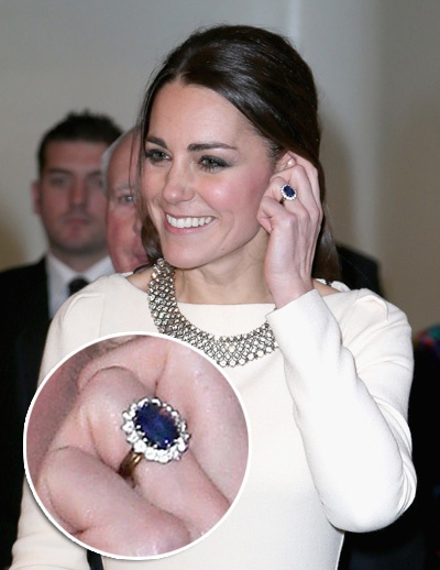 Princesa Kate Middleton, usando um lindo vestido na cor branca, tem sua mão junto ao rosto, onde mostra seu lindo anel de noivado, em safira azul.