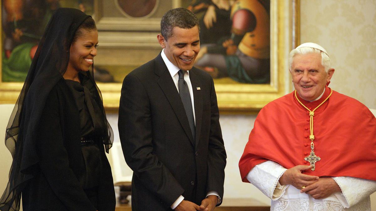 O casal Barack e Michelle Obama, em visita ao Estado do Vaticano, Itália, aparece no Salão Nobre, junto ao Papa Bento XVI. Presidente Obama veste terno na cor preta, camisa branca e gravata cinza escura. Senhora Michelle está usando um vestido, na cor preta e sobre a cabeça usa um manto suave também na cor preta