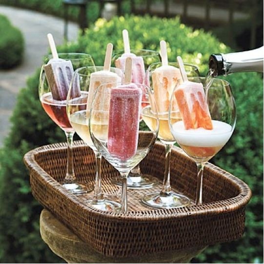 Num jardim, bandeja artesanal, com seis taças de cristal, dentro delas sorvetes coloridos, e está sendo servido champanhe nas taças.