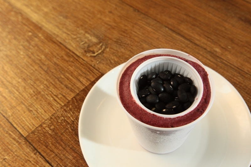 numa mesa de madeira, um copo plástico com uma creme vermelho dentro dele um copinho plástico de café cheio de grãos de feijão.