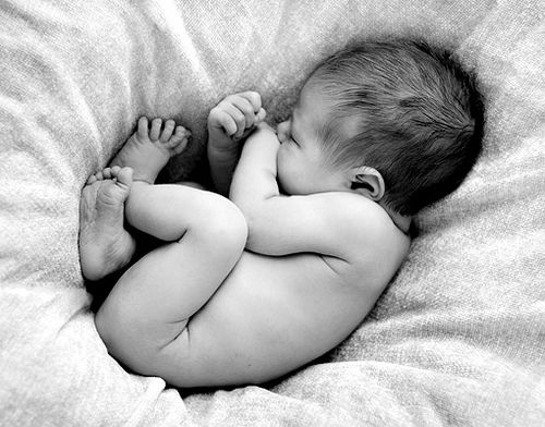 Um bebê recém nascido e peladinho está colocado dormindo sobre lençóis macios. A foto é em preto em branco reforçando a idéia de acolhimento da nova criança.