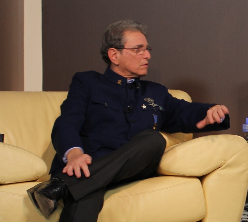 O estilista José Gayegos, de cabelos grisalhos cotados rentes a cabeça, está sentado em um sofá de cor creme vestindo um paletó marinho tipo farda. Na lapela e junto ao ombro usa 4 condecorações