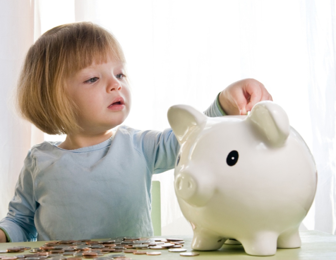 menina de 2 anos, com olhos claros e cabelo loiro curtinho de franjinha, colocando moedas em um cofrinho em forma de porco.