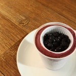 numa mesa de madeira, num prato um copo de plástico com um creme gelado vermelho ,dentro dele um copinho plástico de café com grãos de feijão preto..