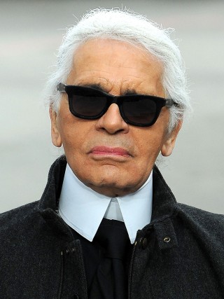Karl Lagerfeld, estilista famoso, sempre com seus cabelos grisalhos, óculos escuros, em foto do rosto, usa camisa branca com golas altas e veste casaco cinza escuro.