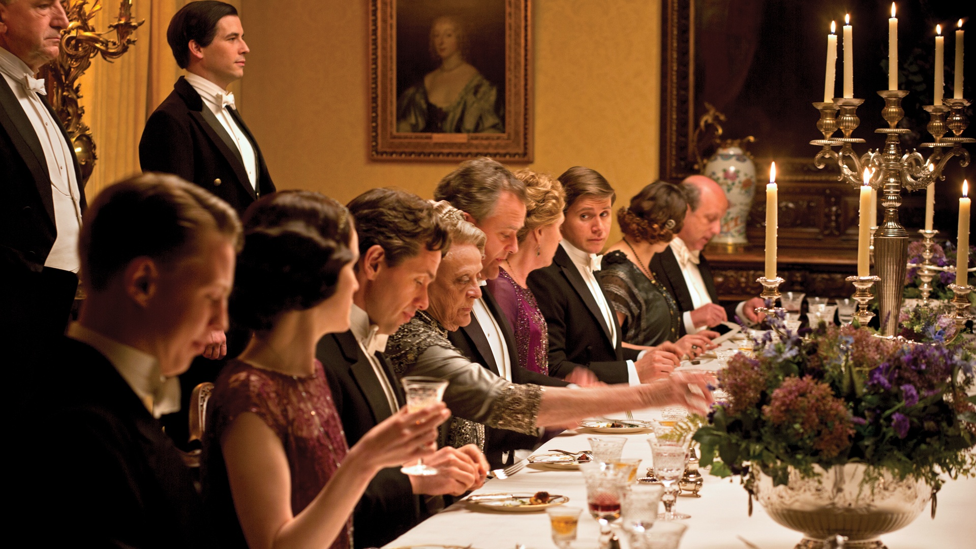 Em uma sala de jantar elegante de um lindo castelo na Inglaterra do início do século XX, os personagens da série inglesa Downton Abbey estão a mesa prontos para jantar. A mesa é oval com uma toalha creme e sobre ela taças de cristal e louça a frente de cada personagem. No centro , candelabros de cinco braços dão um toque final de requinte. O dono da Casa Lorde Crawley está ao centro da mesa e atrás dele o mordomo impecável aguarda ordens em pé, a direita e esquerda de Lorde Crawley, estão sua filha Mary e a sobrinha Rose elegantemente trajadas e a sua frente estão sua mulher, sua terceira filha e o genro, todos concentrados em uma conversa aparentemente séria.