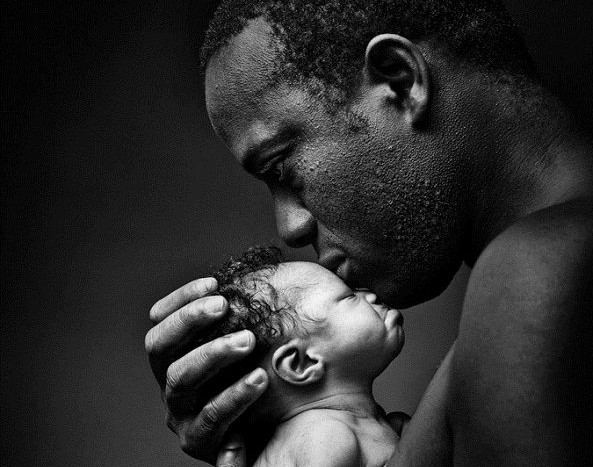 Foto em preto e Branco de um homem negro segurando um recém nascido próximo ao rosto com expressão amorosa. O bebê de olhos ainda fechados está com o rosto encostado no queixo do pai.