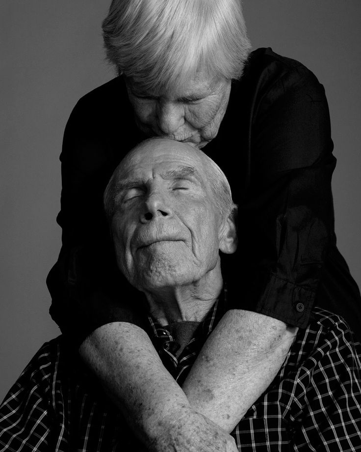 Um casal de senhores de mais de 70 anos se abraça: ele sentado e ela, de cabelos curtos brancos, veste uma malha preta, está em pé e o abraça por trás dando um beijo carinhoso na testa do parceiro que veste uma camisa xadrez. A foto é em preto e branco.