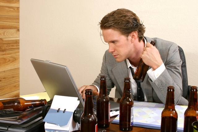 Imagem de um homem usando terno cinza e gravata com o laço solto, está sentado numa mesa de escritório, usando o seu notebook e a sua frente , sobre a mesa temos seis garrafas de cervejas vazias e ele segura mais uma garrafa em sua mão esquerda. Ele olha atentamente para a tela do notebook.