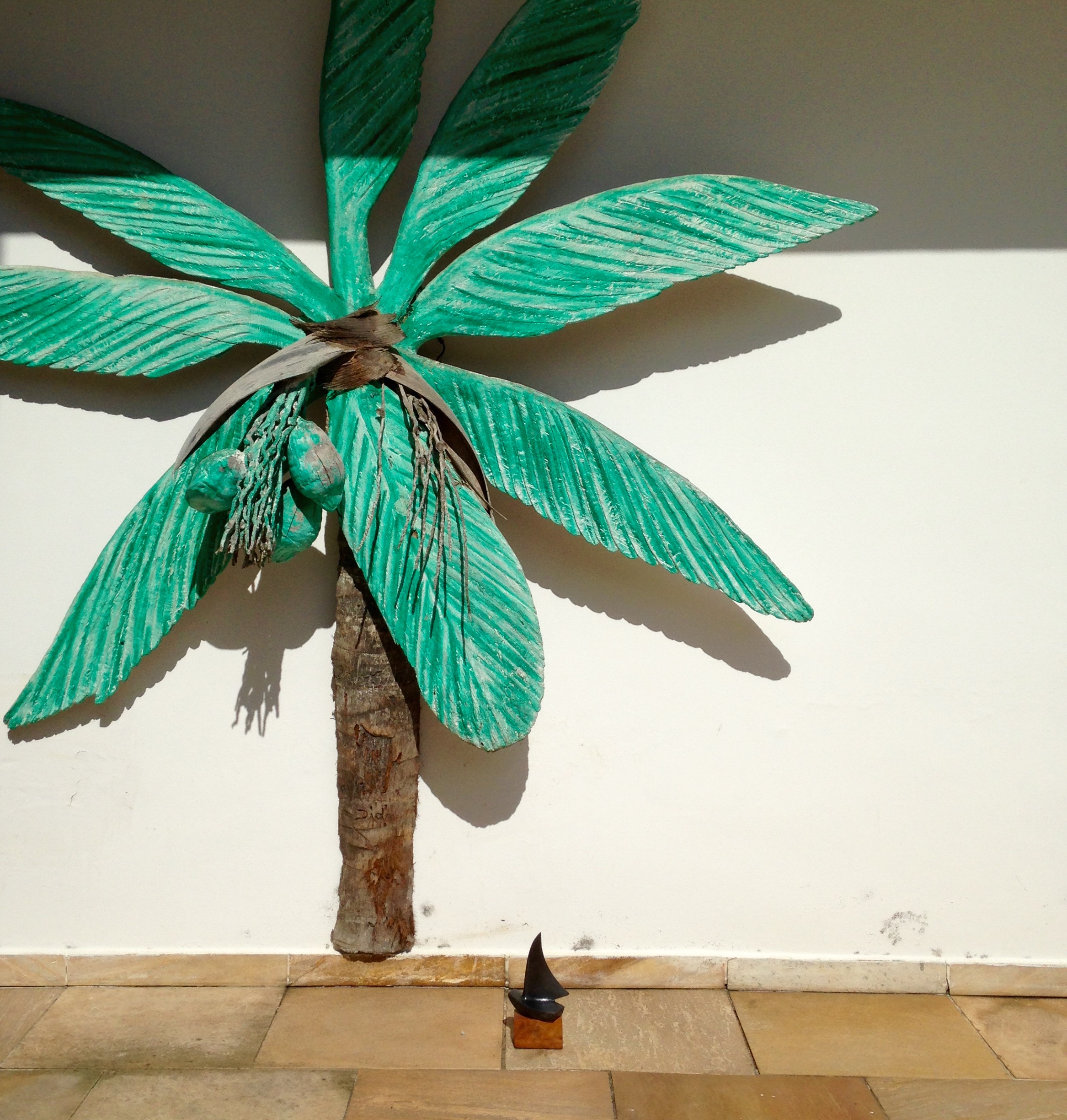 Escultura de Palmeira em madeira colocada junto a parede externa de casa em tamanho natural com o tronco marrom e as folhas verdes