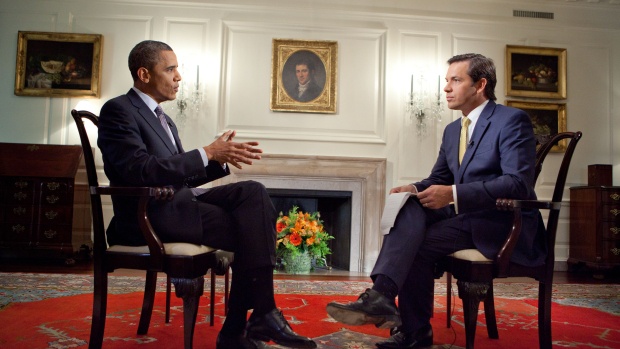 No Gabinete do Presidente, na Casa Branca, nos EUA, o Presidente Barack Obama está sentado em frente a um jornalista, que está realizando uma entrevista. Ambos estão de terno escuro, camisa na cor branca e o Presidente está com uma gravata azul acinzentada e o jornalista com a gravata cor amarela.