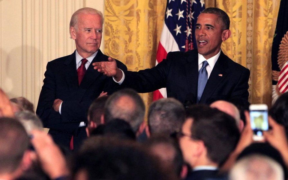 Barack Obama, Presidente dos Estados Unidos ao lado do Vice-Presidente, ambos vestindo terno escuro , camisa branca, Obama com gravata azul e o vice, com gravata na cor vermelha. Ele fala olhando para uma determinada pessoa e gesticula nessa direção, usando o branço direito. Enquanto o Vice-Presidente olha o Obama que está a sua esquerda.