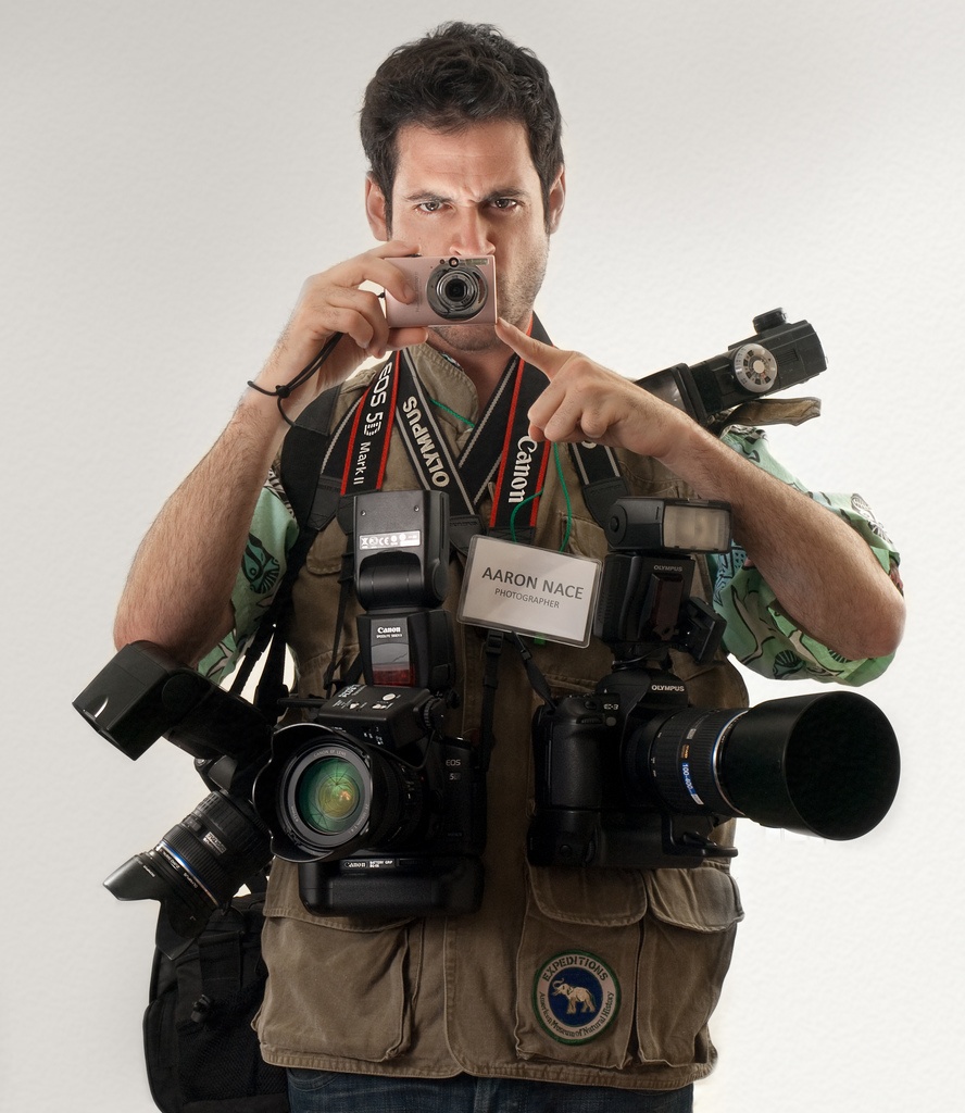 Fotógrafo num imagem frontal , ele alto, cabelos na cor marrom escuro, em sobre o corpo inúmeras câmeras fotográficas de vários tamanhos e lentes de vários tipos.