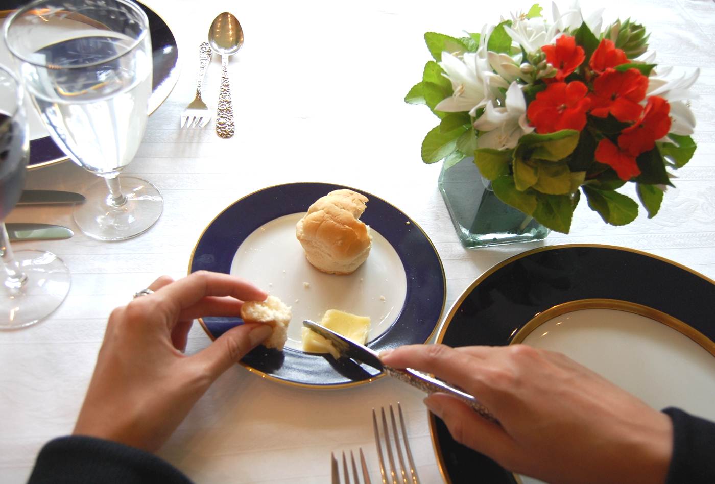 numa mesa de almoço, uma mão segura uma faca e outra segura um pedaço de pão, que está num pratinho. ao lado direito tem um pequeno vaso de flores