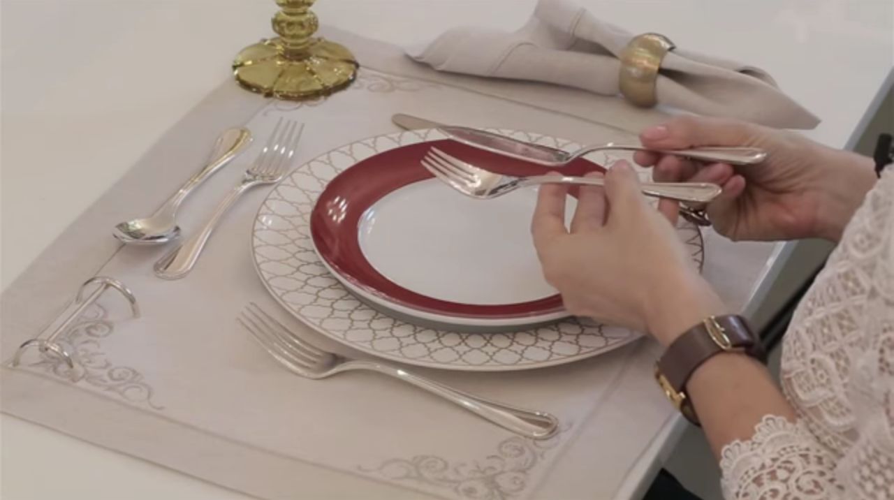 Uma mesa de jantar com toalha cor branca, com prato raso , cor branca, com prato de entrada, com bordas bordô, copos e talheres e copos postados. Uma mão segura um garfo de peixe e uma faca.