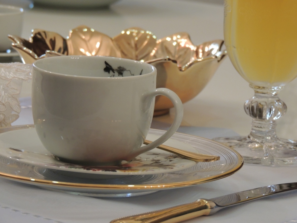 sobre uma mesa, com toalha branca, uma xícara de chá, de porcelana branca, com um detalhe floral na parte interna, sobre um pires com detalhes floral na borda, ao fundo um copo de vidro, com suco de laranja e um pote dourado. Em primeiro plano , uma faca com cabo dourado.