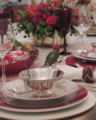 Mesa com pratos de borda vermelho escuro colocada com requinte . Sobre o prato vermelho á outro estampado nos mesmos tons onde está uma taça prateada com abas. Ao fundo um arranjo de rosas vermelhas e em primeiro plano taças de vinho transparentes e de água em tom de rubi. No centro um enfeite de passarinho de porcelana completa a decoração