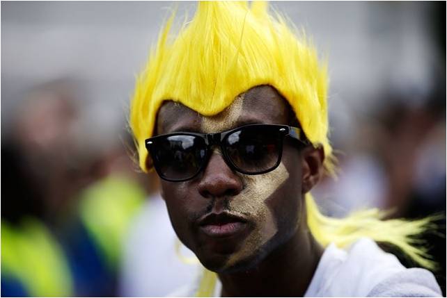 rosto de um homem negro, com o rosto pintado de dourado na lateral esquerda e usa uma peruca arrepiada com cabelos amarelo ovo.