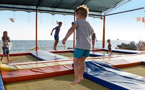Playground a beira mar, onde várias crianças pulam sobre colchões coloridos, em cores vermelho e outros azuis.