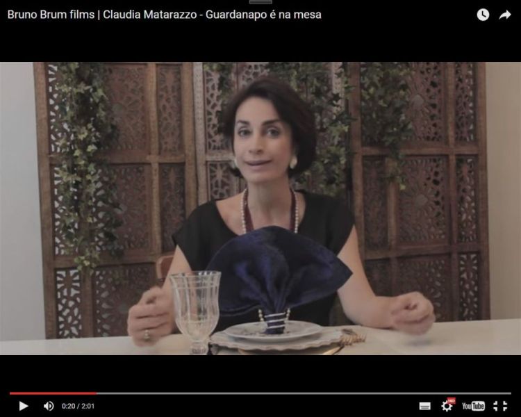 Claudia Matarazzo sentada numa mesa de jantar , diante de um copo com guardanapos escuros a sua frente, ela veste uma blusa cor preta. ao fundo um biombo em madeira.