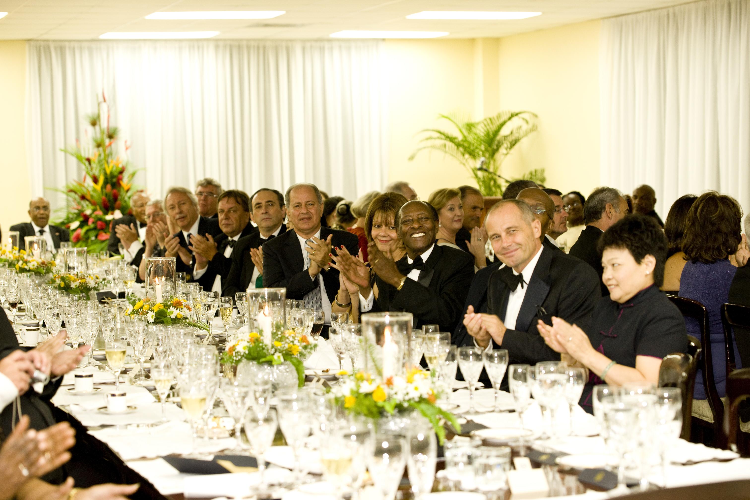 Uma grande mesa de jantar completa, com embaixadores e diplomatas usando black-tie, todos aplaudem.