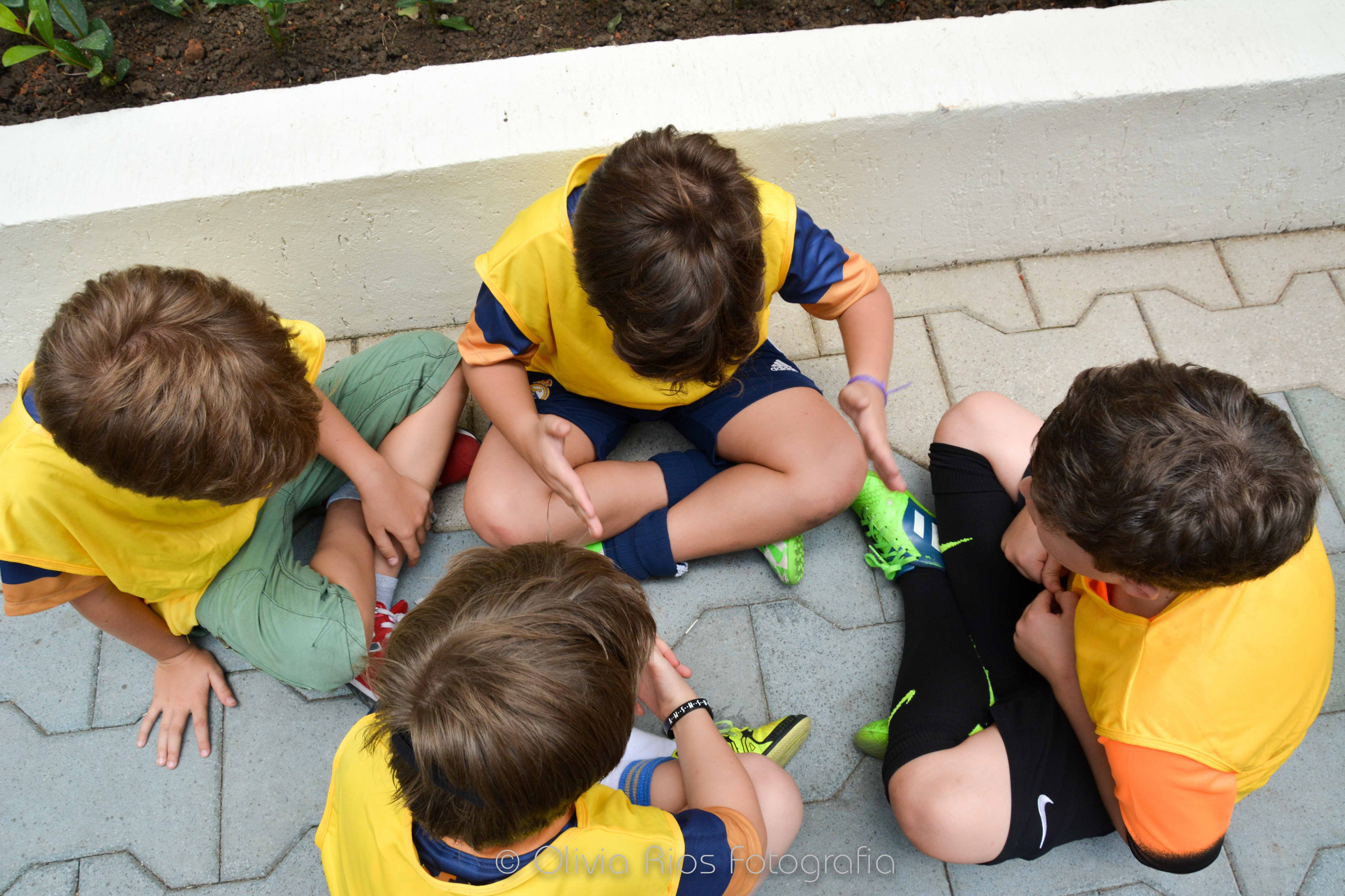 4 crianças sentadas no chão, usando camisetas cor amarela. eles usam chuteiras e diversas cores de shorts. Eles conversam entre si.