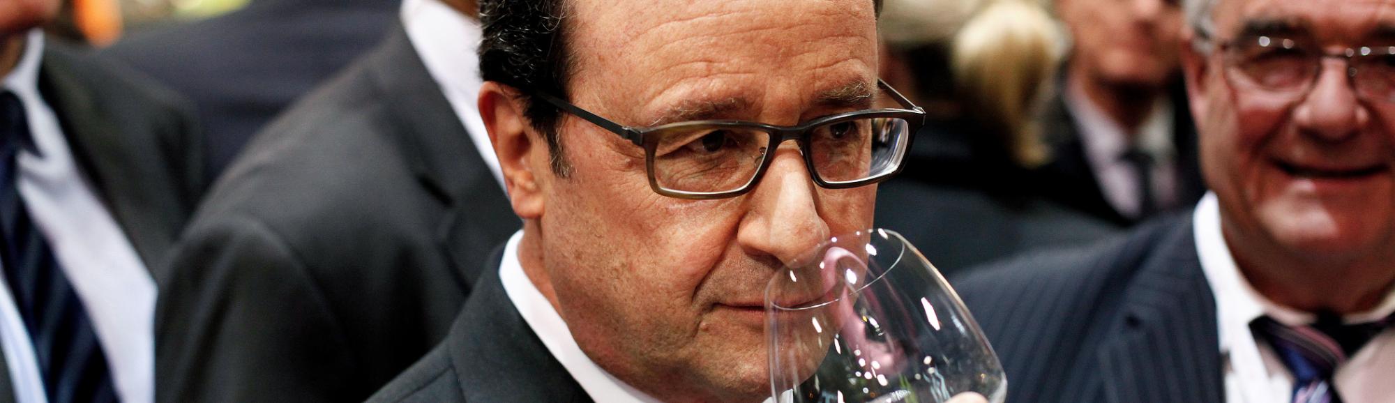 Presidente François Hollande em meio a outras pessoas de terno, ele segura uma taça de vinho junto ao seu nariz,