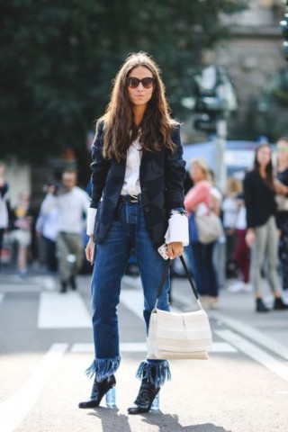 mulher parada numa rua , pousa para foto, usando calça jeans, cor clássica, mas com grande franjas. Usa jaqueta , cor azul marinho escuro, e camisa , cor branca, com mangas extra longas, que produzem algumas dobras junto ao punho. Ela é jovem, usa óculos escuros e tem cabelos longos , castanho escuro. E bota que está usando, tem salto largo e transparente. .
