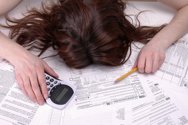 Uma moça está debruçada sobre várias contas em papel com uma calculadora na mão. Só se vê os cabelos da moça e sua postura de desespero.
