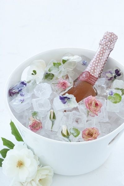 Em um balde de gelo branco que está em um fundo branco se vêem pedras de gelo com flores prensadas com uma garrafa de vinho rosé mergulhada até a metade com o gargalo a vista.