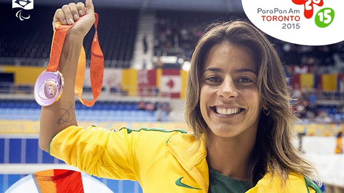 A beira de uma piscina olimpica a nadadora Letícia da seleção brasileira de Natação, ergue seu braço direito que segura nas mãos uma medalha - Ela está sorrindo, cabelos loiros lisos e soltos